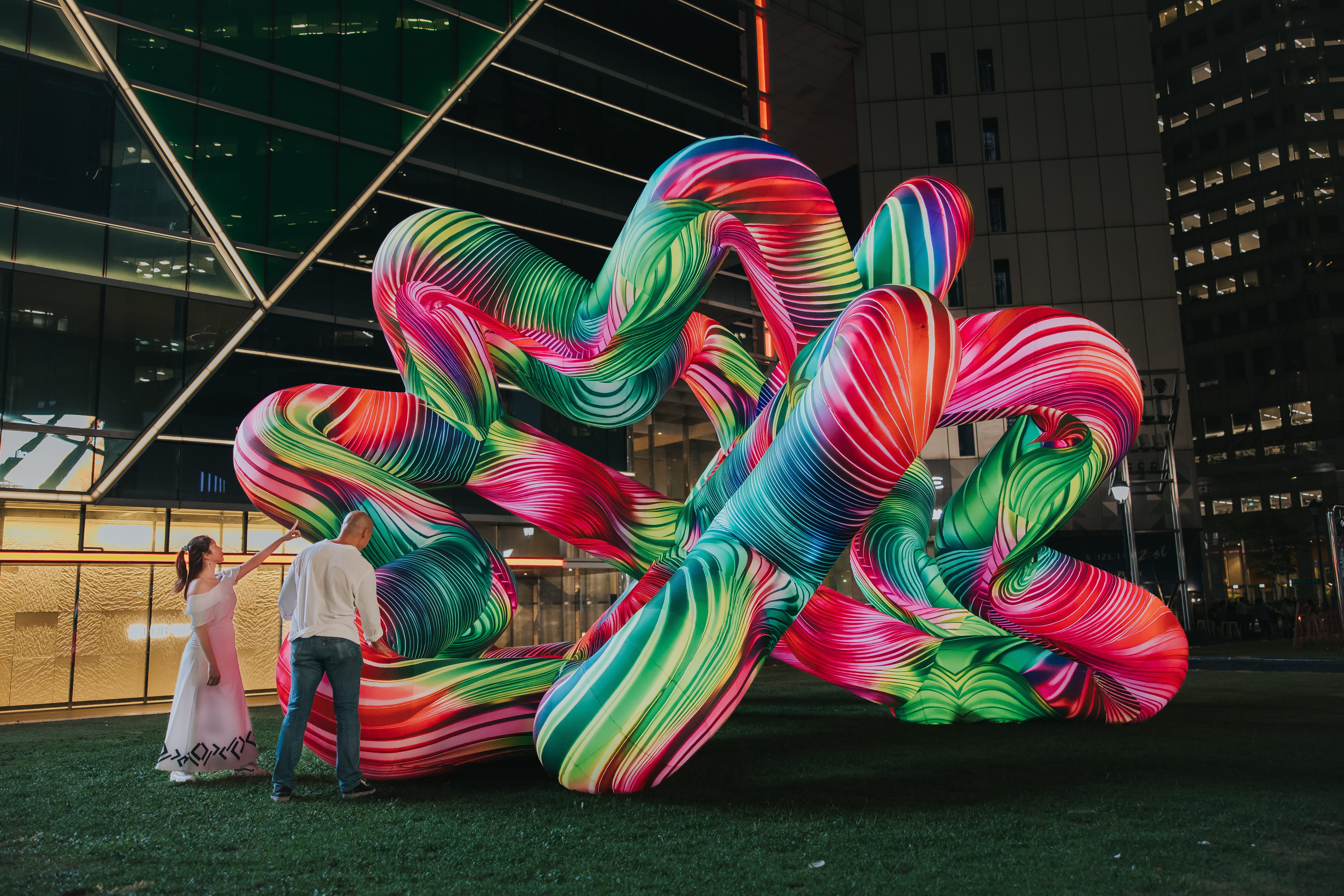 Aerial view of a large, intertwining tubular light sculpture in vivid pink and green, with visitors gathered around it in the dark.